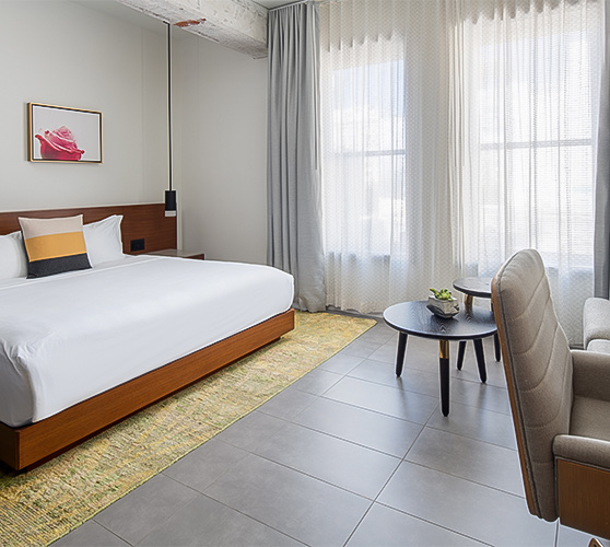 A room with a large bed, a window and some tables with a chair.