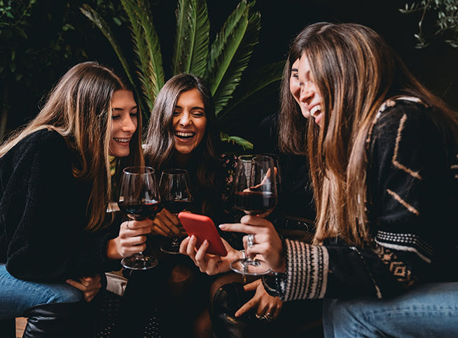 group of girl friends viewing something together on a phone screen while sipping on wine
