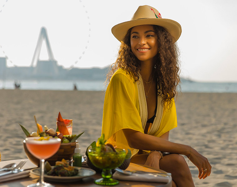 young lady sitting on the beach with a small table with small bites and drinks to enjoy