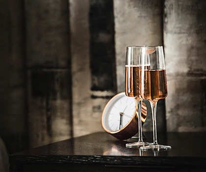 close up of two flutes filled with rose champagne next to a rose gold clock on a black nightstand