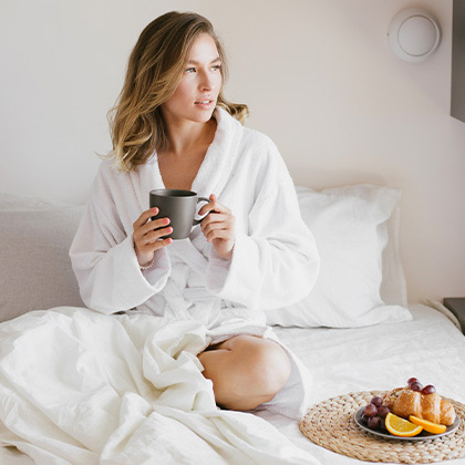 a woman wearing a robe while sitting on a bed drinking coffee next to a small plate of fruit