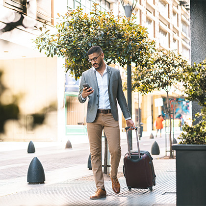 a man wearing business clothes walking down the street with his luggage while looking at his phone